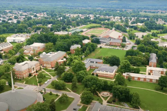 Aerial view of a college campus with multiple brick buildings, green lawns, tree-lined paths, and an athletic track, surrounded by a residential neighborhood and hills—highlighting easy community access to campus facilities.